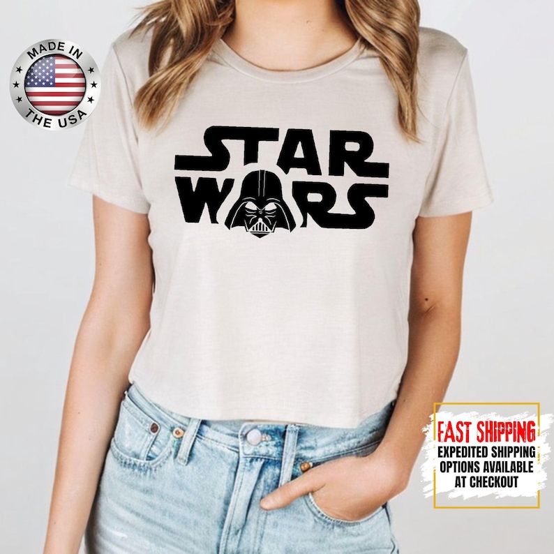 Puede incluir: Camiseta beige claro con el logo negro "STAR WARS" y el casco de Darth Vader. La camiseta est&aacute; hecha en los EE. UU. La modelo lleva vaqueros azules.