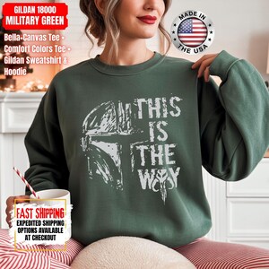 May include: Military green Gildan sweatshirt with the text "THIS IS THE WAY" and a white graphic of a helmet. The sweatshirt also has the text "GILDAN 18000 MILITARY GREEN" and "MADE IN THE USA".