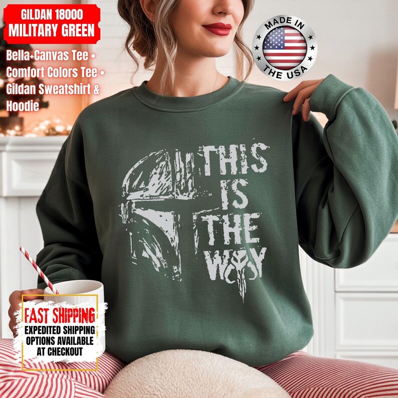 May include: Military green Gildan sweatshirt with the text "THIS IS THE WAY" and a white graphic of a helmet. The sweatshirt also has the text "GILDAN 18000 MILITARY GREEN" and "MADE IN THE USA".
