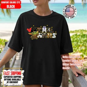 May include: Black t-shirt featuring a Star Wars graphic with Baby Yoda, Mickey Mouse ears, and other characters. The shirt includes the text "STAR WARS" and is made of Comfort Colors material. The shirt also has the text "MADE IN THE USA".