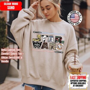 May include: A sand-coloured Gildan 18000 sweatshirt with a colourful Star Wars logo, featuring lightsabers and character-themed letters. The design is reminiscent of a comic book. The collection also includes Bella-Canvas tees, Comfort Colors tees, and hoodies.