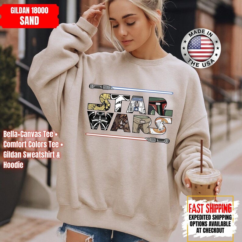 May include: A sand-coloured Gildan 18000 sweatshirt with a colourful Star Wars logo, featuring lightsabers and character-themed letters. The design is reminiscent of a comic book. The collection also includes Bella-Canvas tees, Comfort Colors tees, and hoodies.