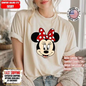 May include: Ivory-colored t-shirt featuring a Minnie Mouse graphic. The design includes Minnie's face with a red bow with white polka dots. The shirt has rolled-up sleeves. Text on the shirt includes "Bella+Canvas Tee", "Comfort Colors Tee", and "Gildan Sweatshirt & Hoodie".