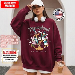 May include: Maroon Disneyland sweatshirt featuring a graphic of Disney characters. The sweatshirt has the text "Disneyland California" and is made of Gildan 18000 material. The image also shows a white baseball cap and headphones.