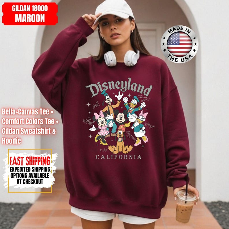 May include: Maroon Disneyland sweatshirt featuring a graphic of Disney characters. The sweatshirt has the text "Disneyland California" and is made of Gildan 18000 material. The image also shows a white baseball cap and headphones.