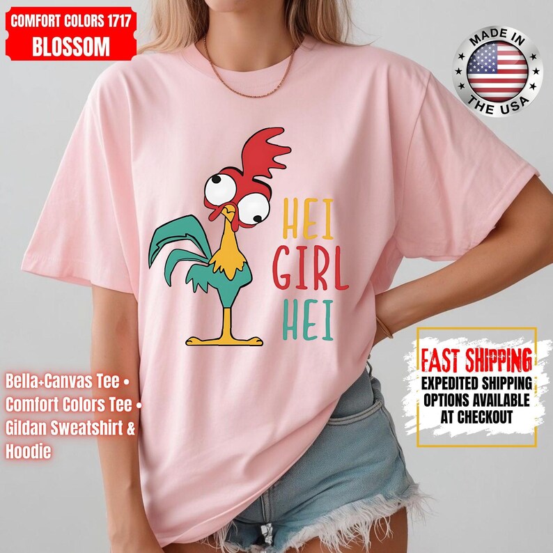 May include: A pale pink t-shirt with a cartoon rooster design and the text "HEI GIRL HEI". The rooster has a red comb, large eyes, and teal feathers. Made in the USA.