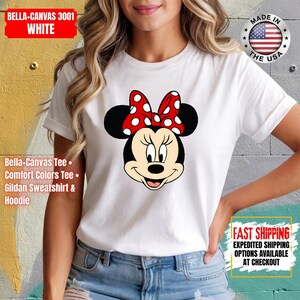 May include: White t-shirt featuring a Minnie Mouse graphic with a red bow with white polka dots. The shirt has the text "BELLA+CANVAS 3001 WHITE" and "MADE IN THE USA". Additional text includes "Bella+Canvas Tee, Comfort Colors Tee, Gildan Sweatshirt & Hoodie".