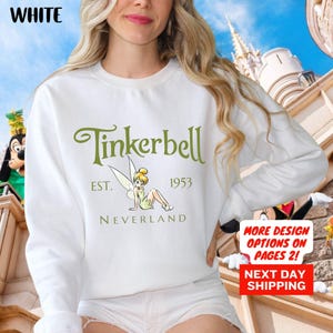 May include: A white long-sleeve sweatshirt featuring a green graphic of Tinkerbell with the text Tinkerbell EST. 1953 NEVERLAND. The shirt includes a white background and long sleeves, showcasing a classic crew neck design with a printed character illustration.