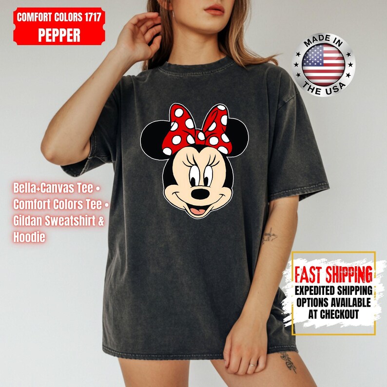 May include: Dark gray Comfort Colors t-shirt featuring a Minnie Mouse graphic with a red bow with white polka dots. The shirt has the text "COMFORT COLORS 1717 PEPPER" and "MADE IN THE USA".