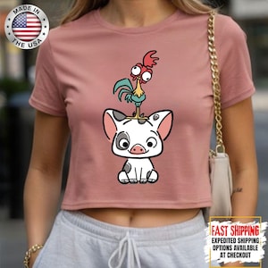 May include: Dusty rose crop top featuring a cartoon illustration of a pig with a rooster on its head. The pig is white with black spots and the rooster has red, blue, and yellow accents. The shirt has a crew neck and short sleeves.