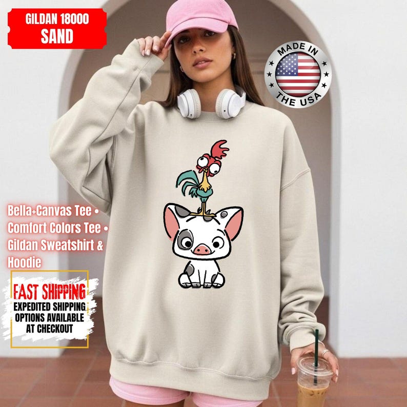 K&ouml;nnte beinhalten: Sandfarbenes Gildan-Sweatshirt mit einem Cartoon-Schwein und einem Hahn-Design. Das Sweatshirt tr&auml;gt den Text "Made in the USA" und "GILDAN 18000 SAND". Das Bild enth&auml;lt auch Text f&uuml;r "Bella-Canvas Tee", "Comfort Colors Tee", "Gildan Sweatshirt & Hoodie" und "FAST SHIPPING".