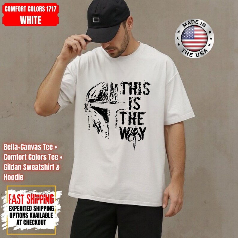 May include: White t-shirt with a black graphic of a helmet and the text "THIS IS THE WAY". The shirt also has a "Made in the USA" emblem. The shirt is a Comfort Colors 1717 Bella-Canvas Tee.