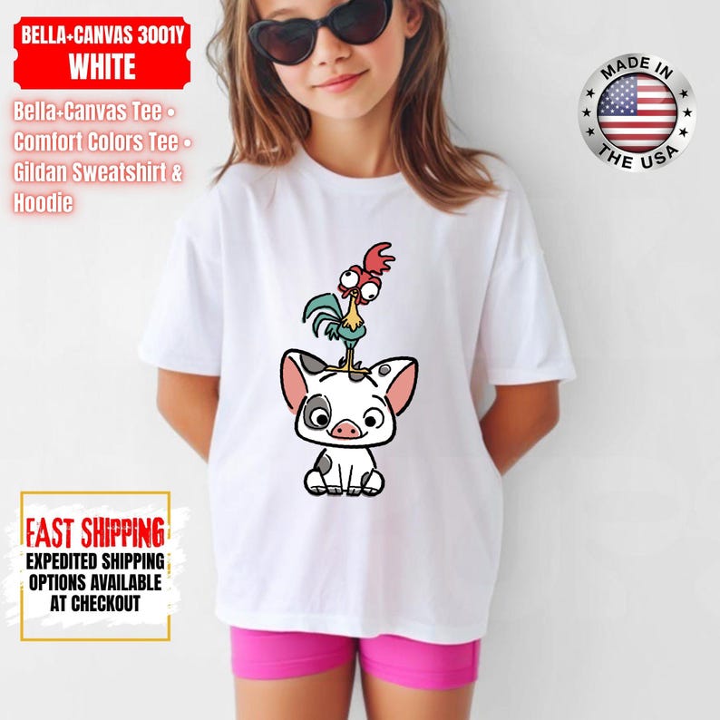 K&ouml;nnte beinhalten: Wei&szlig;es T-Shirt mit einem Cartoon-Schwein und Hahn-Design. Das Shirt tr&auml;gt den Text "BELLA-CANVAS 3001Y WHITE" und "MADE IN THE USA". Es wird mit rosa Shorts getragen. Das Shirt ist Teil der Bella-Canvas Tee, Comfort Colors Tee und Gildan Sweatshirt & Hoodie Kollektion.