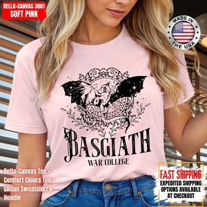 Op de afbeelding: Zachtroze Bella+Canvas T-shirt met een zwarte afbeelding van een draak met uitgespreide vleugels en de tekst "BASGIATH WAR COLLEGE". Het ontwerp bevat sierlijke details en de tekst "MADE IN THE USA".