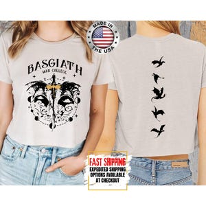 Puede incluir: Un top corto beige claro con un dise&ntilde;o gr&aacute;fico en la parte delantera y trasera. La parte delantera presenta el texto "BASGIATH WAR COLLEGE" con un dise&ntilde;o de espada y alas. La parte trasera tiene una serie de siluetas de dragones negros. Hecho en los EE. UU.