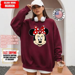 May include: Maroon Gildan 18000 sweatshirt featuring a Minnie Mouse graphic with a red and white polka dot bow. The sweatshirt includes the text "Bella+Canvas Tee", "Comfort Colors Tee", and "Gildan Sweatshirt & Hoodie".