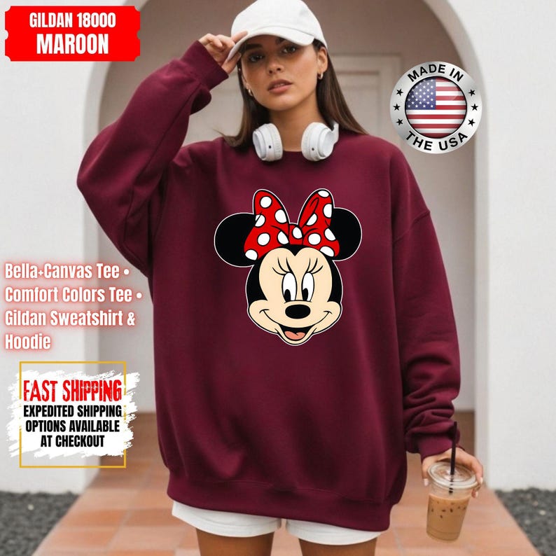 May include: Maroon Gildan 18000 sweatshirt featuring a Minnie Mouse graphic with a red and white polka dot bow. The sweatshirt includes the text "Bella+Canvas Tee", "Comfort Colors Tee", and "Gildan Sweatshirt & Hoodie".