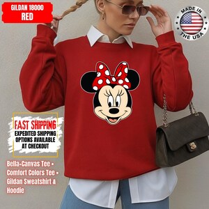 May include: Red Gildan 18000 sweatshirt featuring a Minnie Mouse graphic with a red and white polka dot bow. The sweatshirt has the text "FAST SHIPPING" and "MADE IN THE USA" printed on it. The image also shows a white collared shirt underneath.