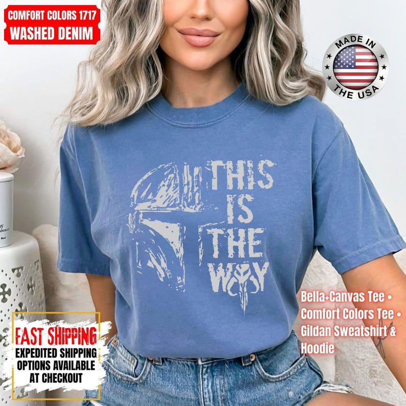 May include: A washed denim blue t-shirt with the phrase "THIS IS THE WAY" in white lettering. The shirt features a graphic of a helmet and a symbol. The shirt is made in the USA.