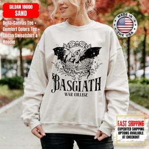 Op de afbeelding: Zandkleurige Gildan-sweatshirt met een zwarte afbeelding van een gevleugeld wezen en de tekst "BASGIATH WAR COLLEGE." De sweatshirt heeft ook de tekst "MADE IN THE USA" en "FAST SHIPPING" erop gedrukt.