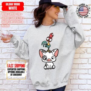 K&ouml;nnte beinhalten: Ein hellgraues Gildan-Sweatshirt mit einem Cartoon-Schwein und Hahn-Design. Das Schwein ist wei&szlig; mit schwarzen Flecken und rosa Ohren, und der Hahn ist gr&uuml;n, rot und gelb. Das Sweatshirt hat auch den Text "GILDAN 18000 WHITE".