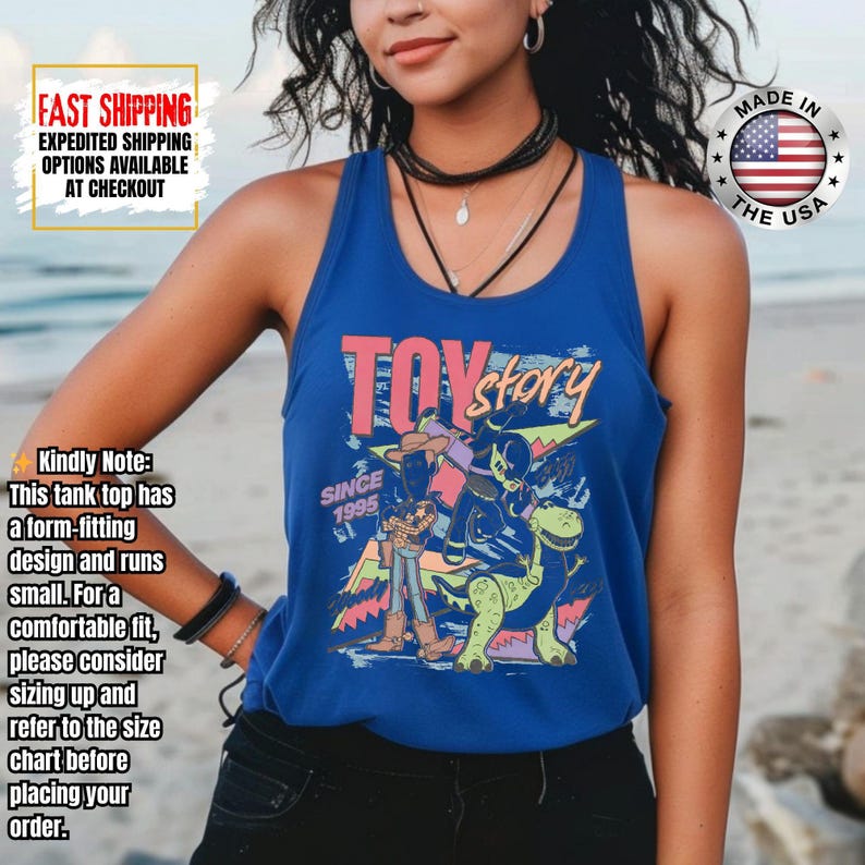 Op de afbeelding: Een blauwe tanktop met de tekst "TOY STORY" in roze en geel, en een afbeelding van personages uit de film. De tanktop heeft ook de tekst "SINCE 1995" en "MADE IN THE USA".