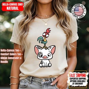K&ouml;nnte beinhalten: Beiges T-Shirt mit einem Cartoon-Schwein und Hahn-Design. Das T-Shirt hat hochgekrempelte &Auml;rmel und den Text "Bella+Canvas 3001 Natural, Bella-Canvas Tee, Comfort Colors Tee, Gildan Sweatshirt & Hoodie". Hergestellt in den USA.
