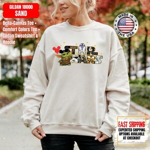 May include: Sand-colored sweatshirt featuring a Star Wars graphic with Baby Yoda holding Mickey Mouse balloons. The text "STAR WARS" is in a block letter style. The sweatshirt has long sleeves and a crew neck. Made in the USA.