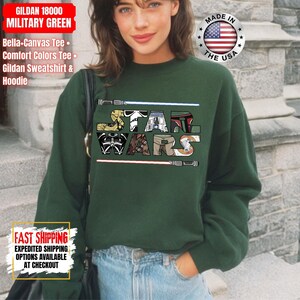 May include: A dark green sweatshirt featuring the text "STAR WARS" in a colourful, stylised font. The design incorporates imagery from the Star Wars franchise. The sweatshirt is a Gildan 18000, Bella+Canvas Tee, Comfort Colors Tee, or Gildan Sweatshirt & Hoodie.