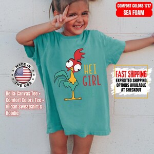 May include: Sea foam green t-shirt with a cartoon rooster and the words "HEI GIRL". The shirt also has the text "MADE IN THE USA" and "Bella+Canvas Tee, Comfort Colors Tee, Gildan Sweatshirt & Hoodie".