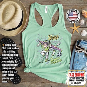 May include: A pale green racerback tank top with a Buzz Lightyear graphic and the phrase "To Infinity and Beyond". The design includes a straw hat, flip-flops, and denim shorts. The tank top is made of a soft material. The image also includes a "Made in the USA" badge.