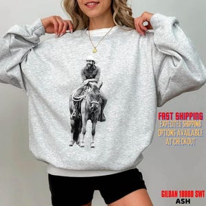May include: A light gray crewneck sweatshirt featuring a black and white graphic print of a cowboy riding a horse. The sweatshirt has long sleeves and a ribbed collar, cuffs, and hem. The graphic is centered on the chest of the garment.