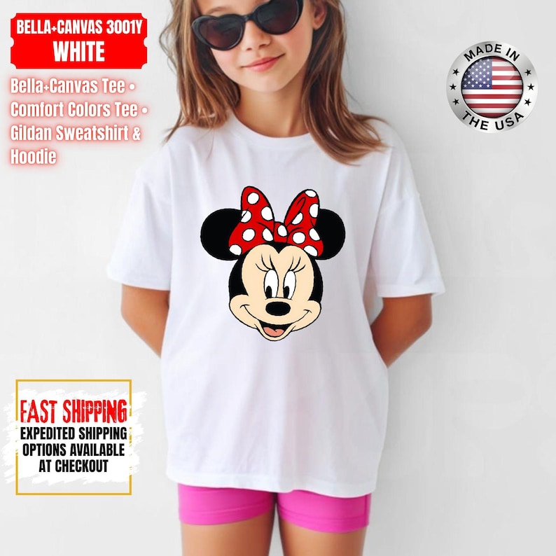 May include: White t-shirt featuring a Minnie Mouse graphic with a red bow with white polka dots. The shirt has the text "BELLA-CANVAS 3001Y WHITE" and "MADE IN THE USA" on it. The shirt is paired with pink shorts.