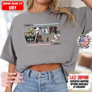 May include: Grey Comfort Colors t-shirt with a colourful "STAR WARS" design. The design incorporates imagery from the Star Wars franchise, with lightsabers above and below the text. Made in the USA.