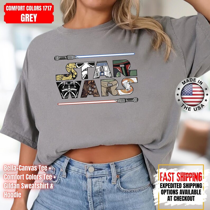 May include: Grey Comfort Colors t-shirt with a colourful "STAR WARS" design. The design incorporates imagery from the Star Wars franchise, with lightsabers above and below the text. Made in the USA.