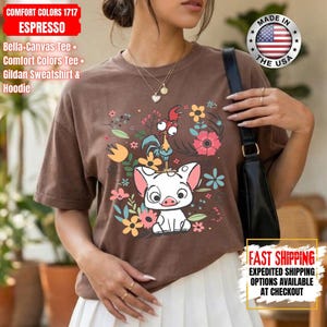 May include: A brown Comfort Colors t-shirt with a cartoon pig and rooster design, surrounded by flowers. The shirt has the text "MADE IN THE USA". It is a Bella-Canvas Tee, Comfort Colors Tee, or Gildan Sweatshirt & Hoodie.