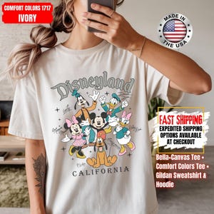 May include: Ivory-colored t-shirt featuring the word "Disneyland" above a graphic of Disney characters including Mickey Mouse, Minnie Mouse, Donald Duck, and Goofy. The shirt also has the text "CALIFORNIA" below the characters.