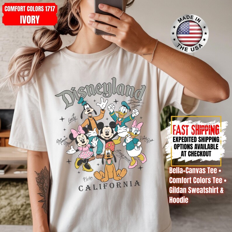 May include: Ivory-colored t-shirt featuring the word "Disneyland" above a graphic of Disney characters including Mickey Mouse, Minnie Mouse, Donald Duck, and Goofy. The shirt also has the text "CALIFORNIA" below the characters.