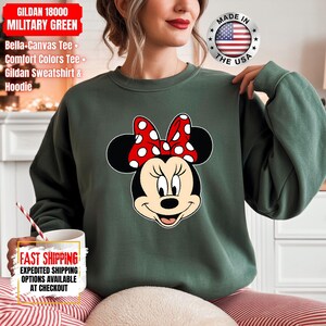 May include: Military green Gildan sweatshirt featuring a Minnie Mouse graphic with a red polka dot bow. The sweatshirt has the text "GILDAN 18000 MILITARY GREEN" and "Bella-Canvas Tee, Comfort Colors Tee, Gildan Sweatshirt & Hoodie". The image also includes a "Made in the USA" emblem.