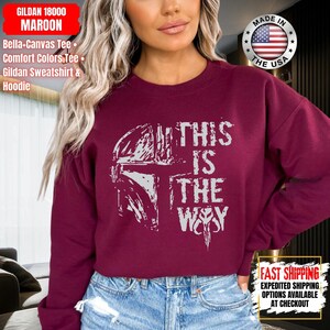 May include: Maroon Gildan sweatshirt with the text "THIS IS THE WAY" in white. The design includes a helmet graphic. The sweatshirt is made in the USA.