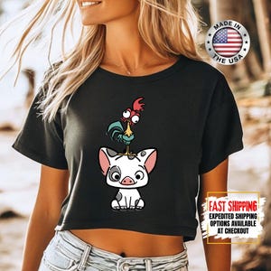 May include: Black crop top featuring a cartoon illustration of a pig with a rooster on top. The pig is white with black spots and pink ears. The rooster has red, blue, and yellow accents. The shirt has a "Made in the USA" graphic.