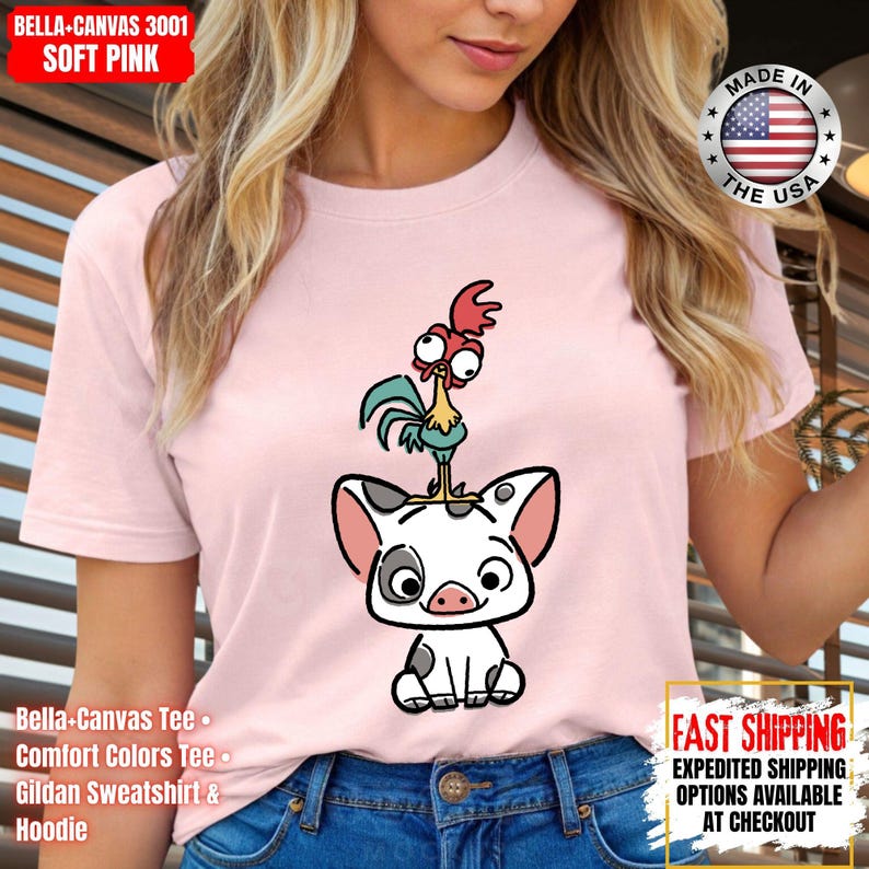 K&ouml;nnte beinhalten: Hellrosa T-Shirt mit einem Cartoon-Schwein und Hahn-Design. Das Shirt tr&auml;gt die Aufschrift "MADE IN THE USA" und "FAST SHIPPING". Das Shirt ist aus weichem Material. Es ist ein Bella+Canvas T-Shirt.