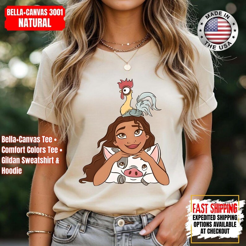 May include: Natural-colored Bella+Canvas 3001 t-shirt featuring a cartoon illustration of a character with long brown hair, a pig, and a rooster. The shirt has rolled-up sleeves. Text on the shirt includes "Bella-Canvas Tee" and "Comfort Colors Tee."