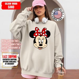May include: Sand-colored Gildan 18000 sweatshirt featuring a Minnie Mouse graphic. The graphic shows Minnie's face with a red polka-dot bow. The sweatshirt has long sleeves and a crew neck. The text "MADE IN THE USA" is also visible.