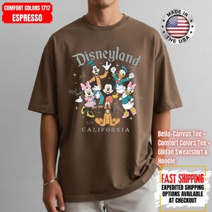 May include: Espresso-colored t-shirt featuring a Disneyland graphic with Mickey Mouse, Minnie Mouse, Goofy, Donald Duck, Daisy Duck, and Pluto. The shirt includes the text "CALIFORNIA" and "MADE IN THE USA".