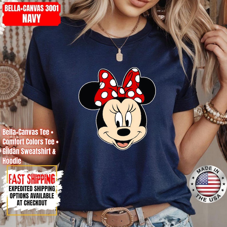 May include: Navy blue Bella+Canvas t-shirt featuring a Minnie Mouse graphic. The graphic shows Minnie Mouse's face with a red bow with white polka dots. The shirt has rolled-up sleeves and is made in the USA.