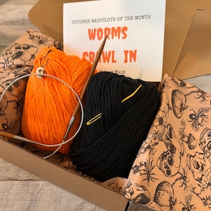 May include: A craft kit in a cardboard box containing one skein of orange yarn, one skein of black yarn, a circular knitting needle, and a small skull stitch marker. A card reads OCTOBER WASHCLOTH OF THE MONTH WORMS CRAWL IN. The box is lined with skull-patterned paper.