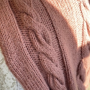May include: A close-up of a soft, pink knitted throw blanket with a cable knit pattern. The blanket is folded and draped over a white surface.