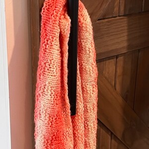 May include: A chunky knit infinity scarf in shades of coral and cream hangs on a black metal rod. The scarf's texture is visible, with a repeating pattern. The background features a wooden door.