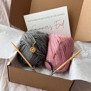 May include: A knitting kit in a cardboard box containing one skein of grey yarn with a sunflower stitch marker, one skein of pink yarn, and circular knitting needles. A card reads SEPTEMBER WASHCLOTH OF THE MONTH Summer's End.